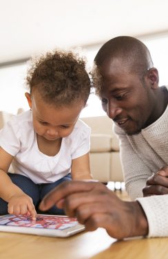 Young afro-american father at home with his cute little daughter lying on the floor watching something or playing a game on tablet.